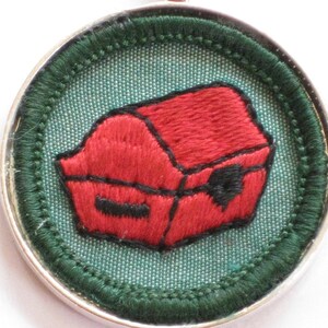 May include: A red embroidered treasure chest pendant on a green fabric background with a silver metal frame.