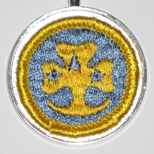 May include: A silver pendant necklace with a round, blue and yellow embroidered design. The design features a yellow cross with three stars.
