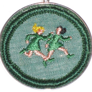 May include: A silver pendant necklace with a round green fabric charm featuring an embroidered image of two girls in green dresses running. The charm is surrounded by a green embroidered border.