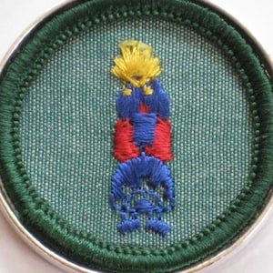 May include: A green and silver Girl Scout badge with a colorful embroidered design of a torch with flames.