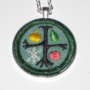 May include: A silver necklace with a round green pendant featuring an embroidered design of a tree with four seasons: a yellow apple, a green leaf, a white snowflake, and a red leaf.