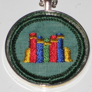 May include: A green fabric pendant with a colorful embroidered design of stacked blocks. The pendant is framed in a silver circle.