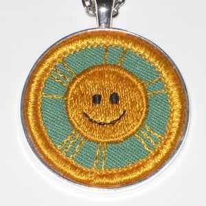 May include: A gold pendant with a green and yellow embroidered smiley face design. The pendant is round and has a silver frame.