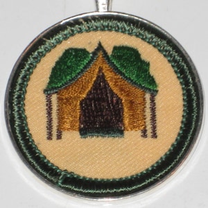 May include: A round, green and gold embroidered patch with a tent design. The tent is brown with a dark brown door and green flaps. The patch is surrounded by a green border.