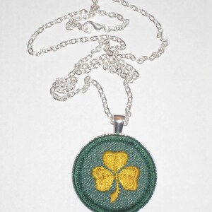 May include: A silver chain necklace with a round pendant featuring a green shamrock with a gold embroidered design.