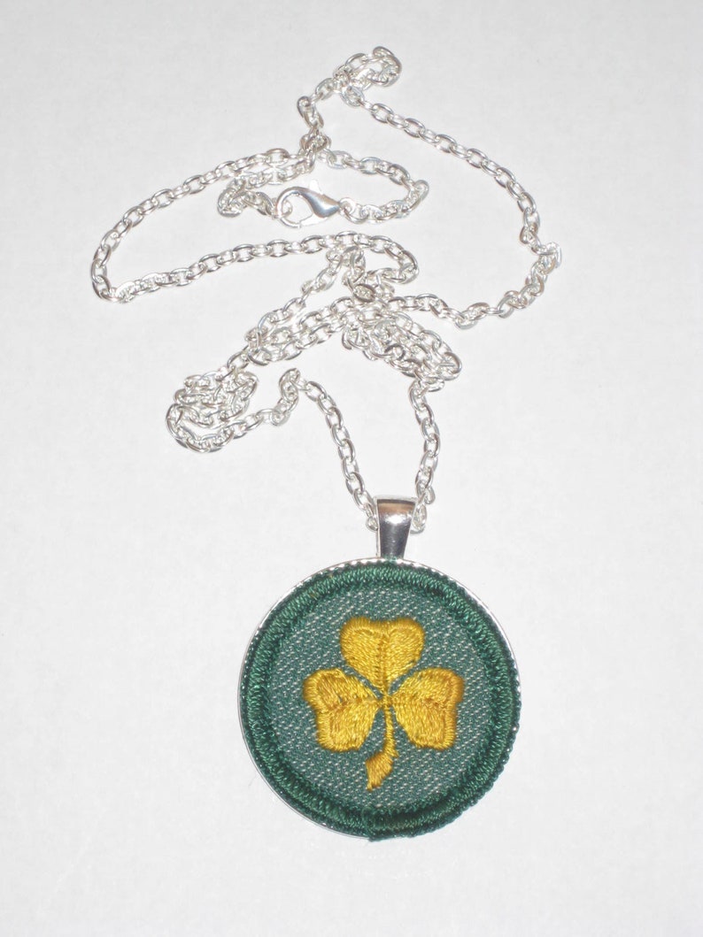 May include: A silver chain necklace with a round pendant featuring a green shamrock with a gold embroidered design.