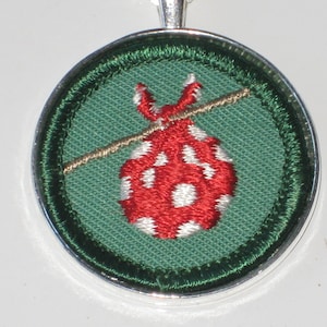 May include: A green and red embroidered patch with a red and white polka dot bag being pulled by a string. The patch is surrounded by a silver metal frame.