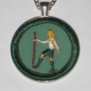 May include: A green and silver Boy Scout pendant with a white embroidered figure holding a staff.