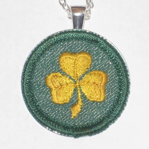 May include: A silver chain necklace with a round pendant featuring an embroidered green shamrock with a yellow center.
