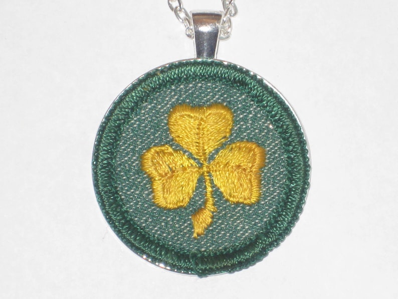 May include: A silver chain necklace with a round pendant featuring an embroidered green shamrock with a yellow center.