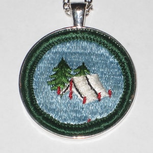 May include: A round, green and blue embroidered patch with a white tent and two green trees. The patch is attached to a silver chain.