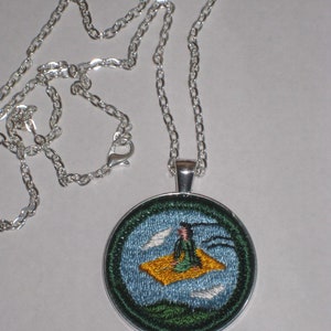 May include: A silver chain necklace with a round pendant featuring an embroidered image of a person sitting on a flying carpet. The carpet is yellow and the person is wearing a green robe. The background is blue with white clouds.