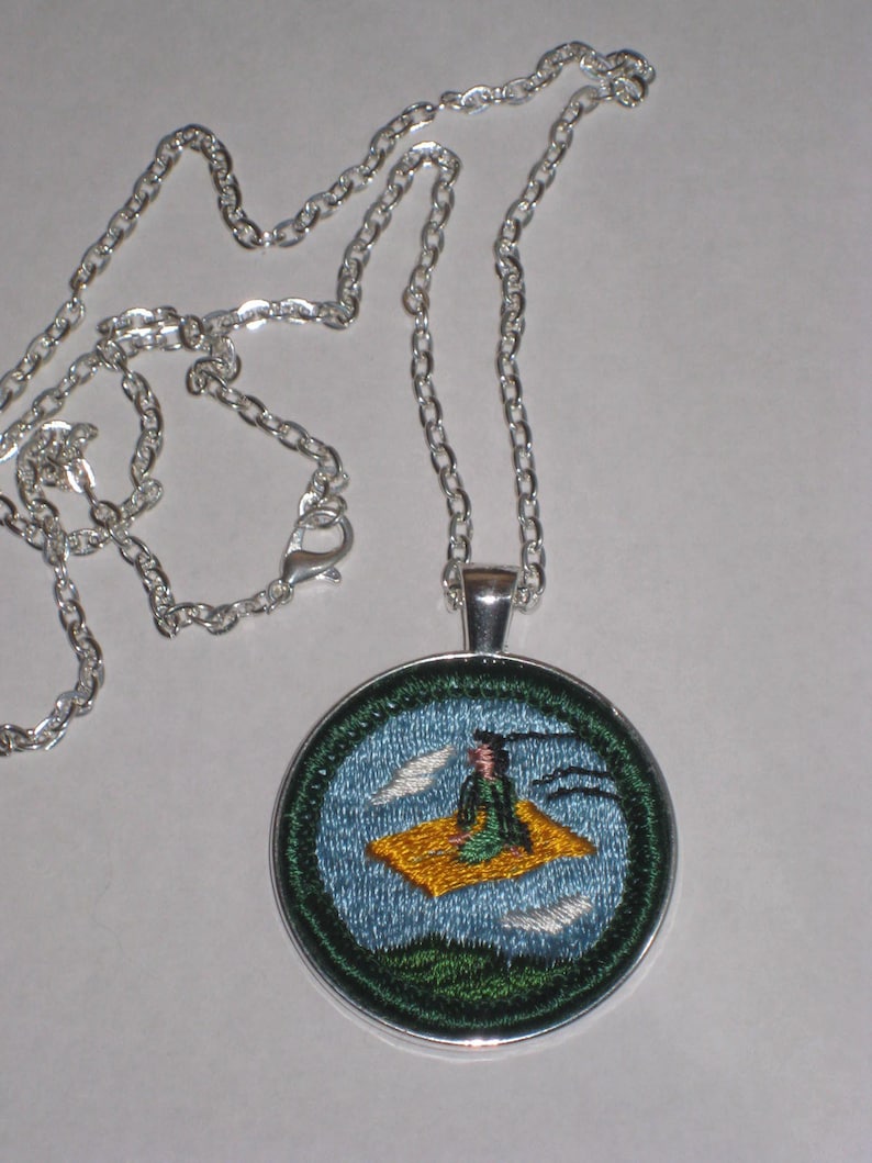 May include: A silver chain necklace with a round pendant featuring an embroidered image of a person sitting on a flying carpet. The carpet is yellow and the person is wearing a green robe. The background is blue with white clouds.