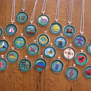 May include: Twenty-two round, green, embroidered patches with silver chains. The patches feature various designs, including a compass, a sailboat, a tent, a musical note, a campfire, a horse, and a key.