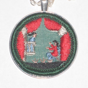 May include: A round, silver-toned pendant with a green, red, and white embroidered design. The design depicts two figures in a scene with a red curtain.