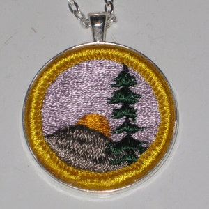 May include: A silver pendant necklace with a round, embroidered patch. The patch features a purple sky with a yellow sun setting behind a gray mountain and a green pine tree.