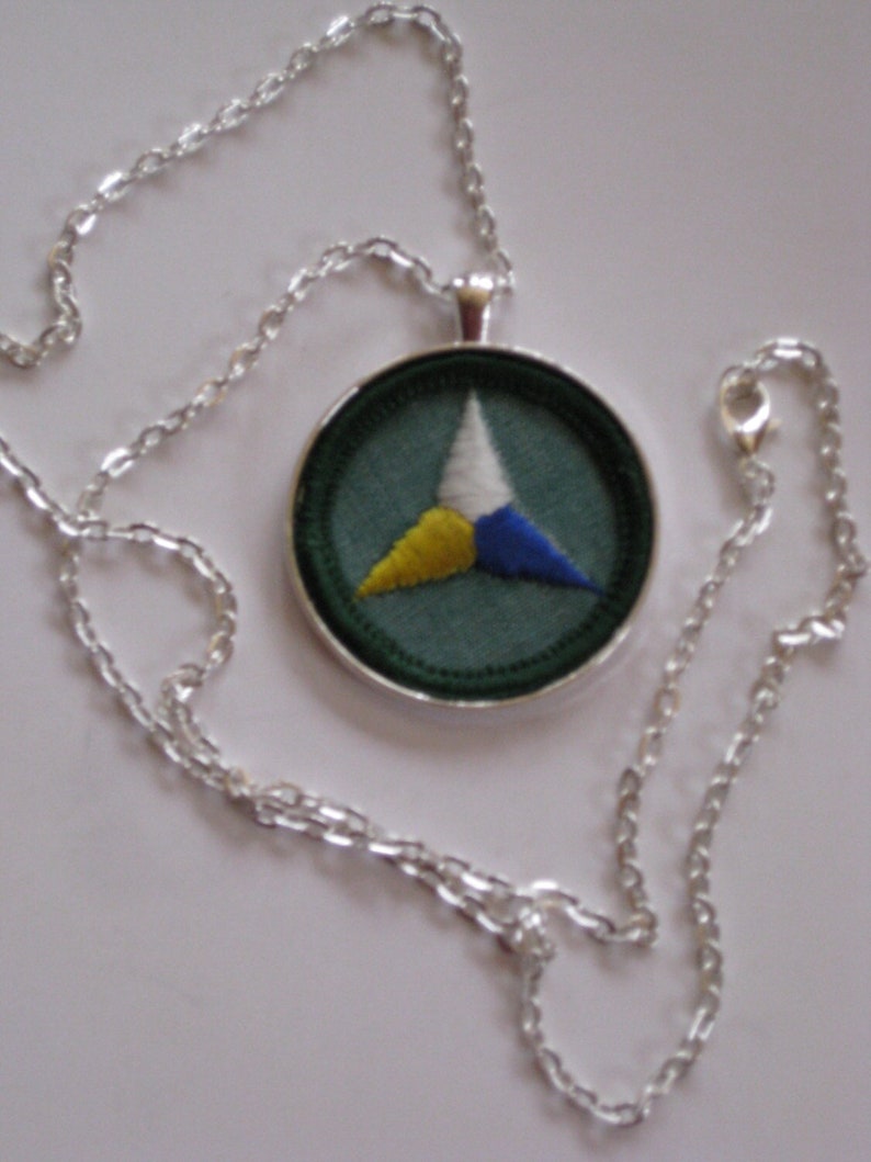 Puede incluir: Un collar de cadena de plata con un colgante redondo con fondo verde y un tri&aacute;ngulo blanco, amarillo y azul.