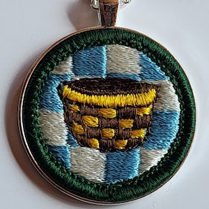 May include: A round, silver-toned pendant with a green border and a blue and white checkered background. A brown basket with yellow accents is embroidered in the center.