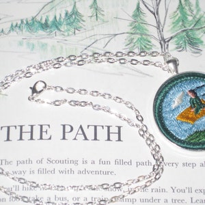 May include: A silver chain necklace with a round pendant featuring an embroidered image of a person flying on a yellow cloud. The pendant is blue with green accents and white clouds. The text "THE PATH" is visible in the background.