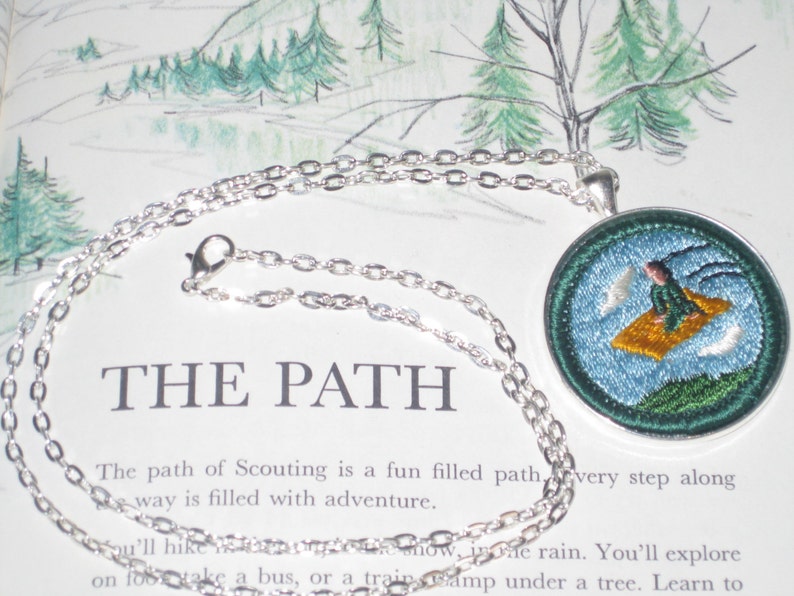 May include: A silver chain necklace with a round pendant featuring an embroidered image of a person flying on a yellow cloud. The pendant is blue with green accents and white clouds. The text "THE PATH" is visible in the background.