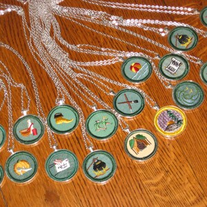 May include: A collection of Girl Guide badges on silver chains. The badges are round and feature various designs, including a tent, a campfire, a first aid kit, and a compass. The badges are all green with white and yellow accents.