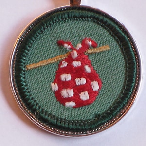 May include: A green and silver round pin with a red and white embroidered design of a bag with a stick going through it.