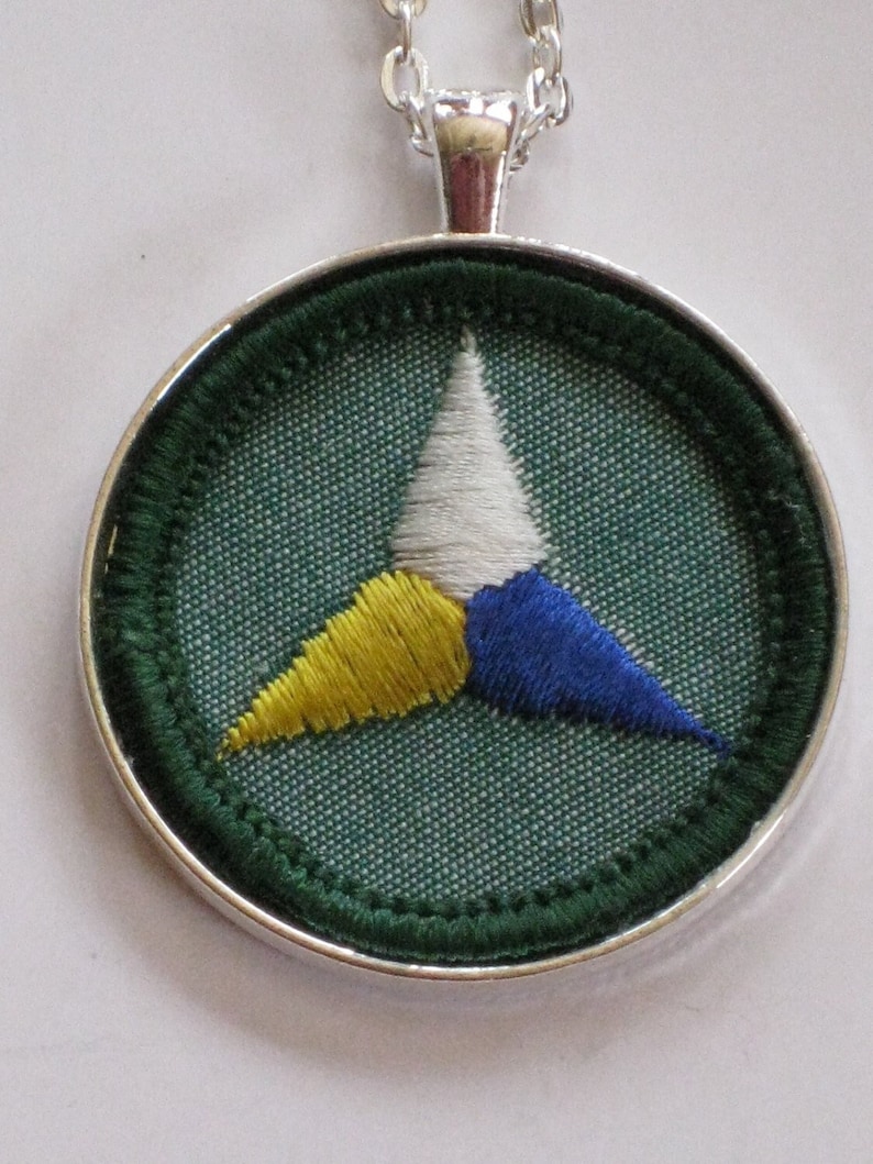 Puede incluir: Un collar con un colgante de plata y un c&iacute;rculo de tela verde con un tri&aacute;ngulo bordado en blanco, amarillo y azul.