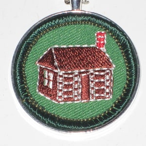 May include: A round, silver-colored pendant with a green fabric background. The pendant features an embroidered image of a brown cabin with a red chimney.