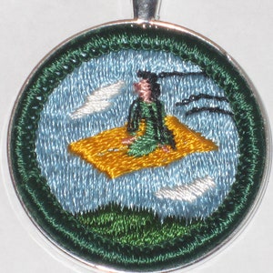 May include: A silver pendant featuring an embroidered design of a person sitting on a cloud with birds flying around them. The design is in shades of green, blue and yellow.