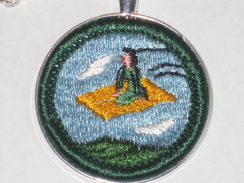 May include: A silver pendant featuring an embroidered design of a person sitting on a cloud with birds flying around them. The design is in shades of green, blue and yellow.