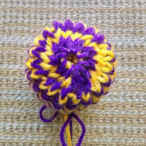 May include: A handmade, round, crocheted item in a vibrant combination of yellow and purple. The yarn is tightly woven, creating a textured surface. A loop of purple and yellow yarn extends from the base. The item is likely a decorative pom-pom or similar craft.
