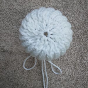 May include: A white crocheted sphere with a textured, ribbed surface pattern radiating from a central opening. Two long, thin white yarn strands extend downwards from the base of the sphere, creating a simple, dangling loop design.