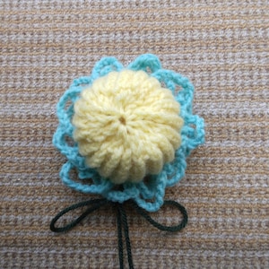 Peut inclure: Une fleur crocheté avec un centre jaune et une couche extérieure bleu clair. La fleur est attachée à une tige verte.