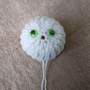 May include: A round, white crocheted sphere featuring two bright green plastic eyes with black pupils. The white yarn is worked in a textured, ribbed pattern that radiates from the center. Two long, thin white yarn strands hang from the bottom.