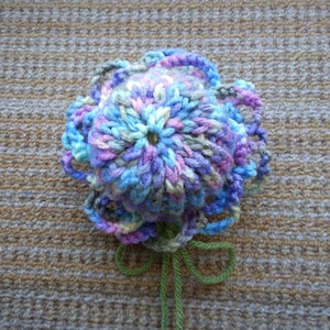 May include: A crocheted flower with a blue, purple and pink colour scheme. The flower has a green stem.