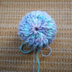 May include: A blue and purple yarn pom-pom with a hole in the center. The yarn is tied together with a blue and turquoise string.