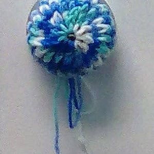 May include: A blue, white, and green crocheted flower with a beaded center and a long blue and white string hanging from the bottom.