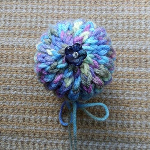 May include: A blue, green and purple yarn pom-pom with a blue flower-shaped button in the centre.