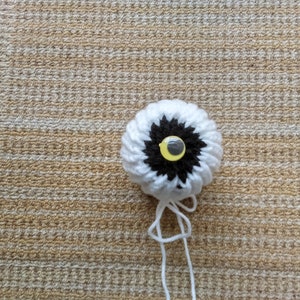 Peut inclure: Un œil fait à la main au crochet avec des détails blancs, noirs et jaunes. L'œil a un anneau extérieur blanc, un centre noir et une pupille jaune et noire. Du fil blanc s'étend du bas de l'œil. L'œil est sur une surface texturée et beige.