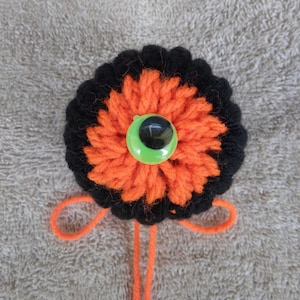 May include: A circular crochet ornament featuring a vibrant orange center surrounded by a thick black border. A single large, bright green plastic eye with a black pupil is centered on the orange yarn. Two orange yarn loops extend from the bottom.