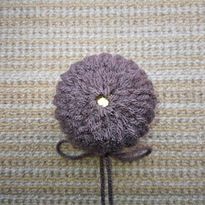 May include: A brown knitted pom-pom with a gold button in the center. The pom-pom is attached to two long brown strings.
