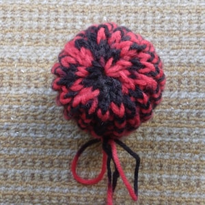 May include: A handmade, round pom-pom in red and black yarn. The pom-pom is knitted with a textured pattern and has loose strands of red and black yarn hanging from the bottom. The background is a textured, tan surface.