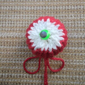 May include: A handmade crochet flower with a red base, white petals, and a green and gray button center. The flower has a red yarn stem with a loop at the end. The craft item is displayed on a textured, tan surface.