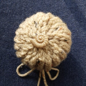 May include: A beige knitted item with a gold spiral embellishment. The item is handmade and has a textured appearance. It is tied with a matching string. The background is a dark blue fabric. The item is likely a decorative element.