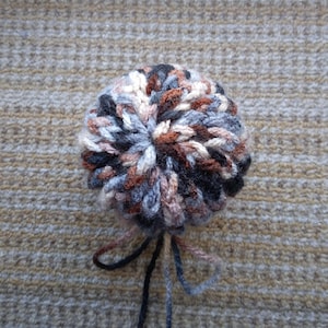 May include: A multi-colored yarn pom-pom with black, brown, white, and gray yarn. The pom-pom is tied with the yarn ends.