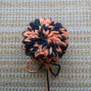 May include: A handmade, round pom-pom in shades of orange and navy blue yarn. The pom-pom is centered in the image, with two strands of yarn extending downward. The background is a textured, tan surface.