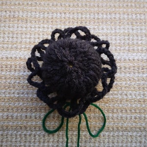 May include: A black crocheted flower with a green yarn stem. The flower is made with a thick yarn and has a textured, fluffy appearance.