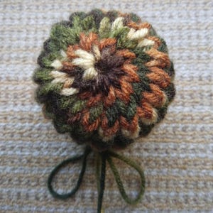 Peut inclure: Un objet rond fait à la main au crochet, avec un motif camouflage vert, marron et beige. L'objet a une ficelle vert foncé nouée en nœud à la base. La texture du fil est visible.