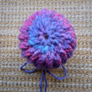 May include: A handmade, crocheted flower in shades of purple, pink, and blue. The yarn is tightly woven, creating a textured, round shape. A length of blue yarn extends from the base. This item is suitable for crafts and decorations.