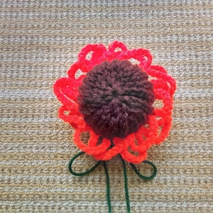 May include: A handcrafted crochet flower featuring a dark brown central part and vibrant red petals. Green yarn creates the stem and leaves. The flower is placed on a textured, beige background.